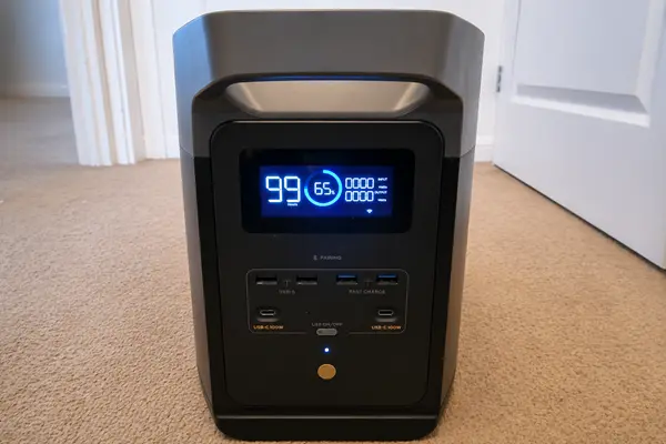 A front view of an EcoFlow Delta 2 portable power station showing the illuminated digital display, USB ports, and power button while sitting on a carpeted floor.